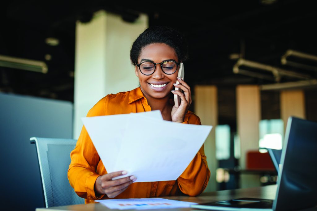 Happy black businesswoman talking on cellphone and checking documents in office, working with papers at workplace